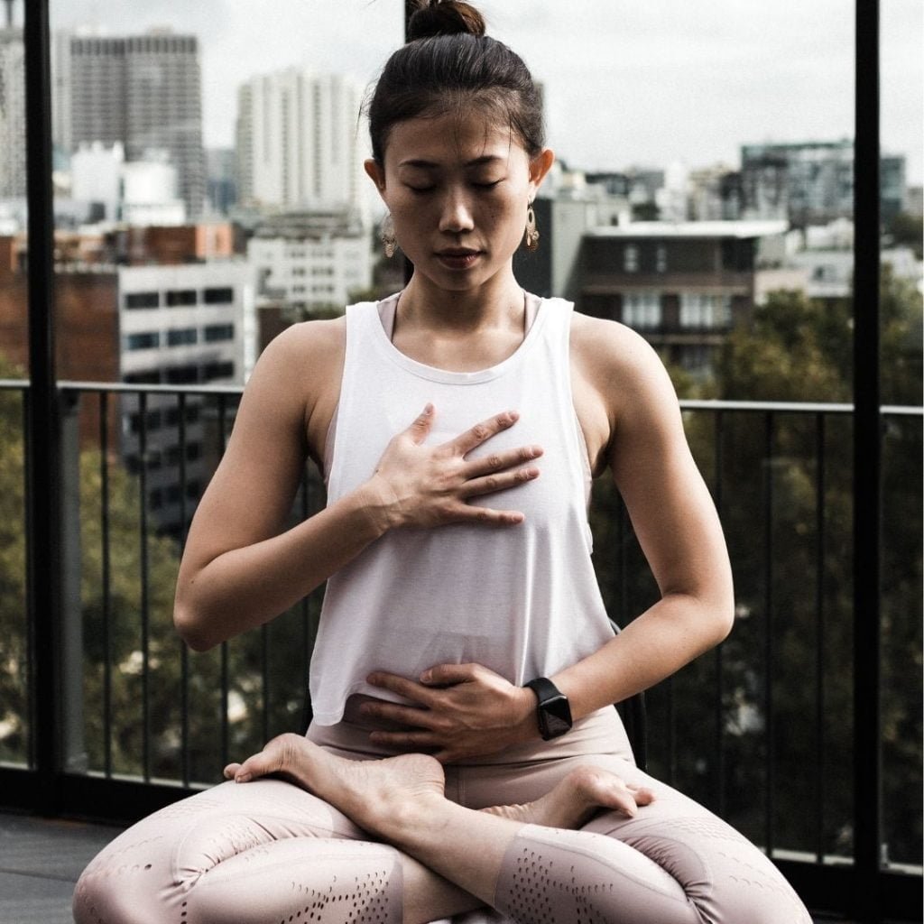 woman sitting on a terrace or balcony with city buildings in the background. The woman is sitting cross-legged with her eyes closed -- one hand is on her chest and one on her abdomen