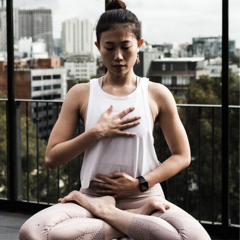 woman sitting on a terrace or balcony with city buildings in the background. The woman is sitting cross-legged with her eyes closed -- one hand is on her chest and one on her abdomen
