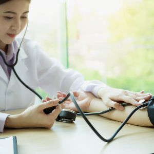 Doctor in white coat with stethoscope in ears (and on a patient's arm) looking at blood pressure reading