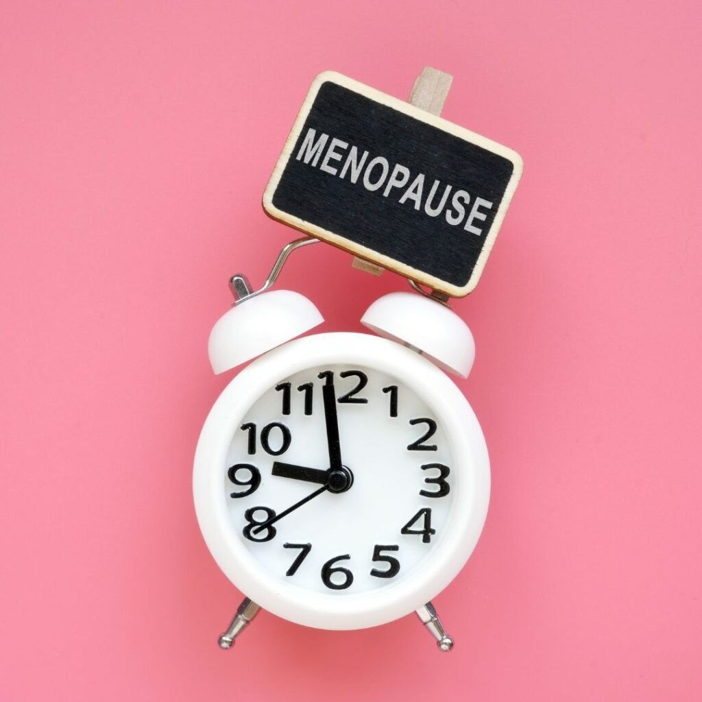Pink background. In the foreground is an analogue white alarm clock with a small chalkboard clip that says: MENOPAUSE
