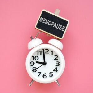 Pink background. In the foreground is an analogue white alarm clock with a small chalkboard clip that says: MENOPAUSE