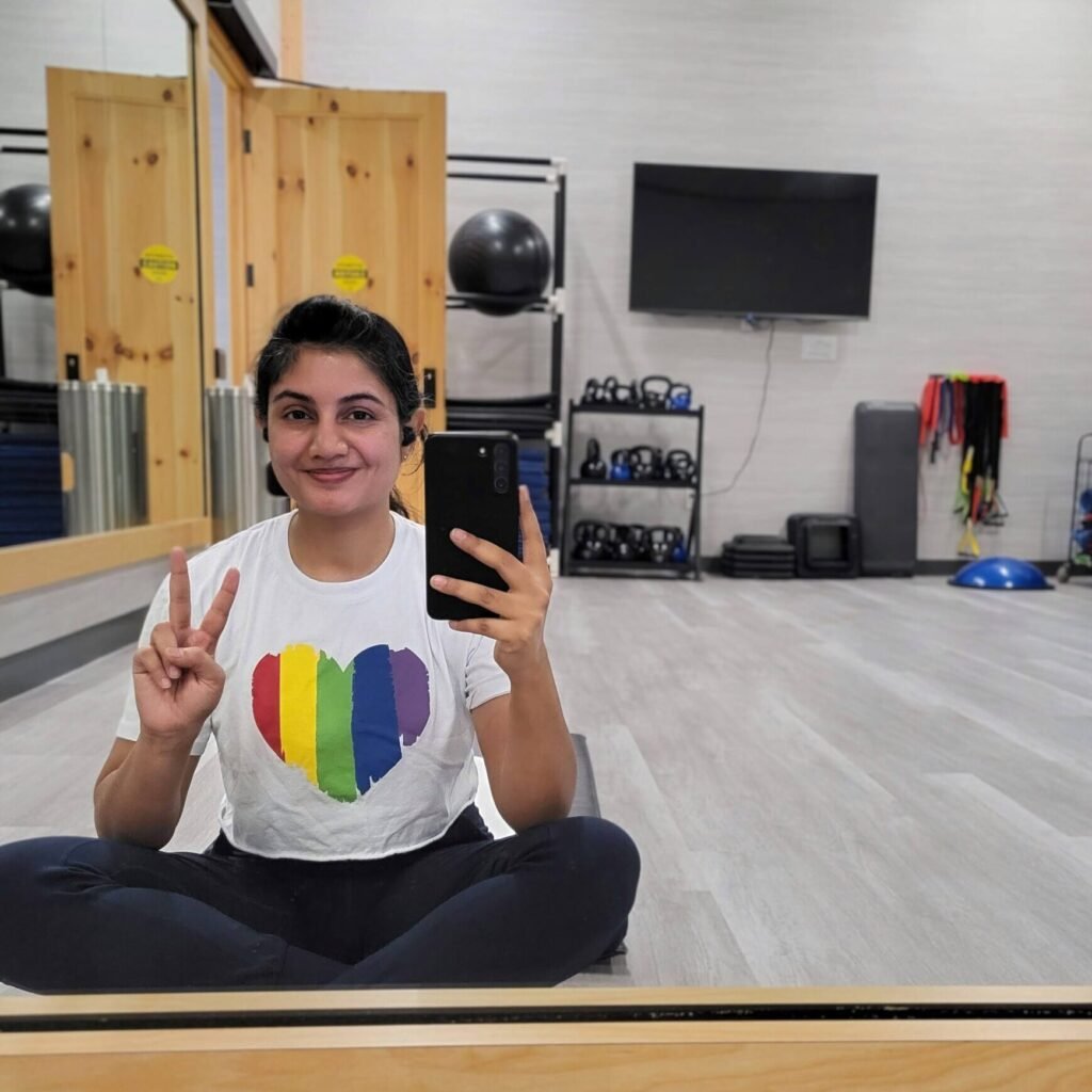 brown-skinned woman, Dr. Anne, reflected in a mirror wearing a white shirt with a rainbow heart on it, sitting in a gym room with exercise equipment in the background.