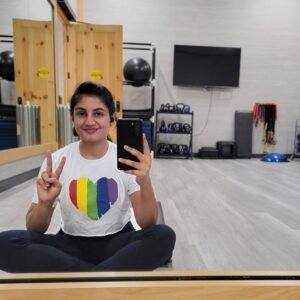 brown-skinned woman, Dr. Anne, reflected in a mirror wearing a white shirt with a rainbow heart on it, sitting in a gym room with exercise equipment in the background.