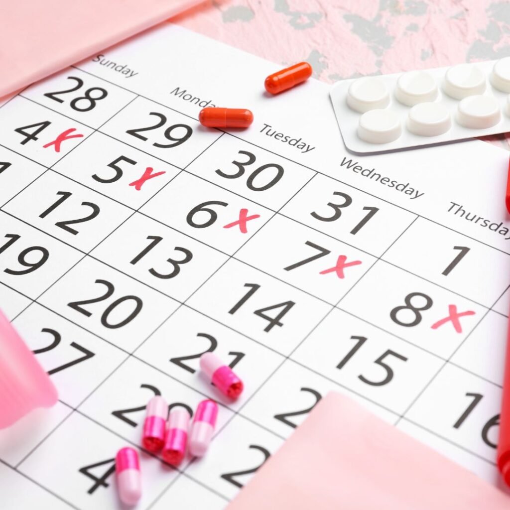 Picture with an offset calendar in white with Xs on 5 days in red denoting a period. There are pink sticky notes, red and pink capsules, a pink menstrual cup, and a blister pack on the calendar.
